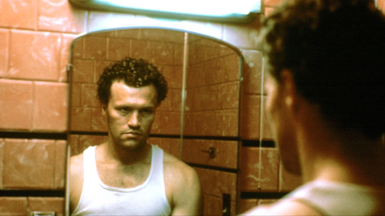 A man in a white undershirt stares into a grimy mirror.