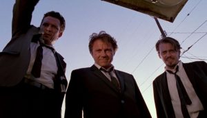 Three men in black suits, white shirts and black ties peer down into an open car trunk