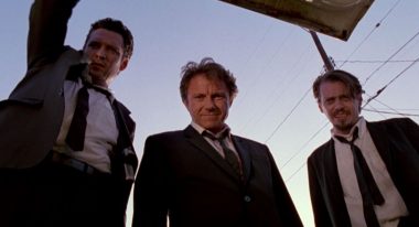 Three men in black suits, white shirts and black ties peer down into an open car trunk