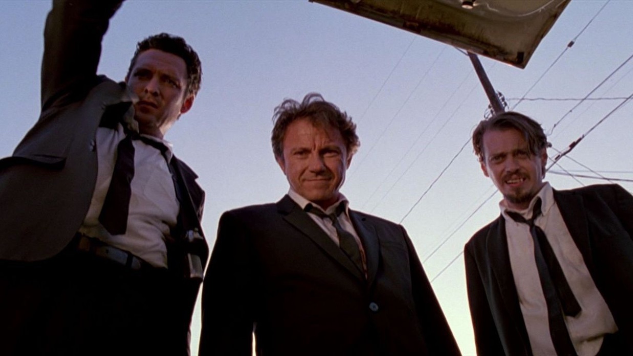 Three men in black suits, white shirts and black ties peer down into an open car trunk