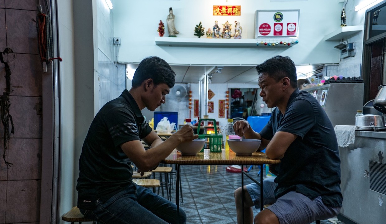 A young man sits across from an older man at a bare-bones Taiwanese noodle shop. They are eating in tandem.