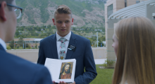 A young man with blond hair in a blue suit showing a picture of Jesus Christ to a a couple with their backs to the camera