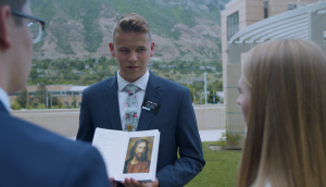 A young man with blond hair in a blue suit showing a picture of Jesus Christ to a a couple with their backs to the camera