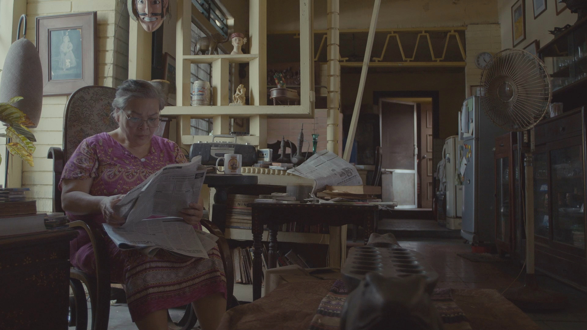 An older woman sitting on a chair, reading a newspaper, in a living room.