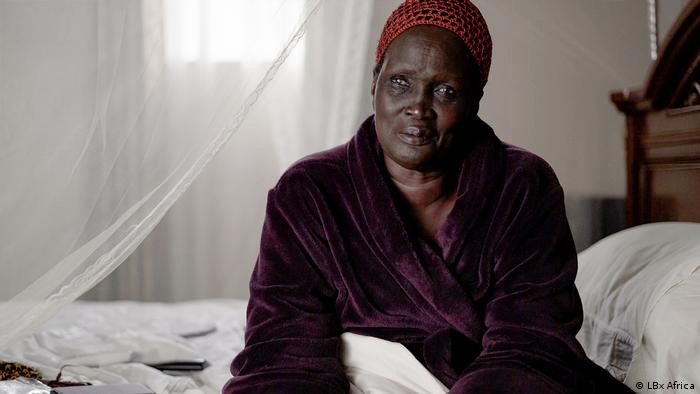 A middle-aged Black woman in a mauve bathrobe and red headscarf on a white bedspread