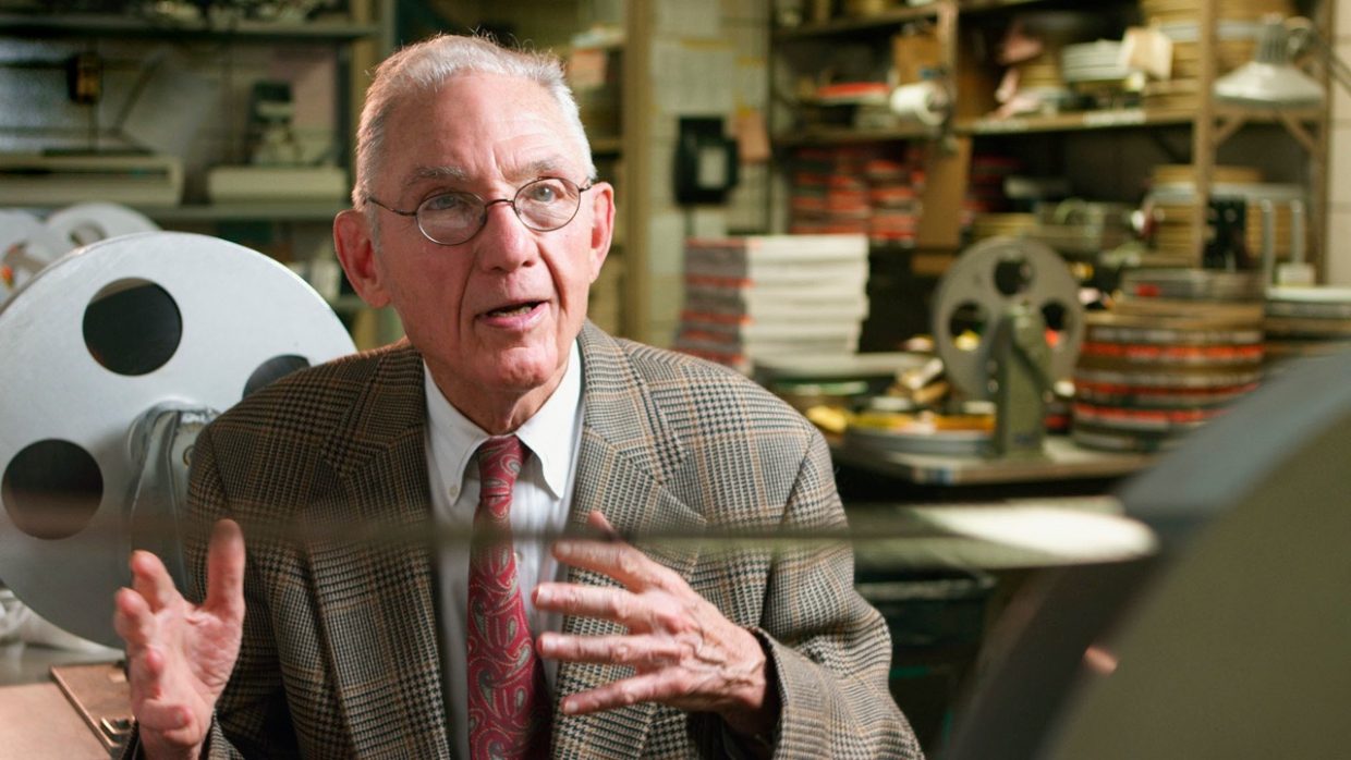 An old white man with glasses and a light brown suit in front of film reels at a film laboratory