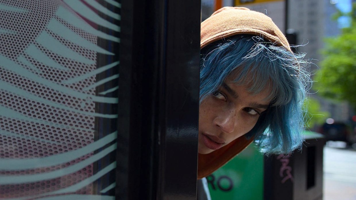 A Black woman with blue hair and an tan hat peering around the corner of a building