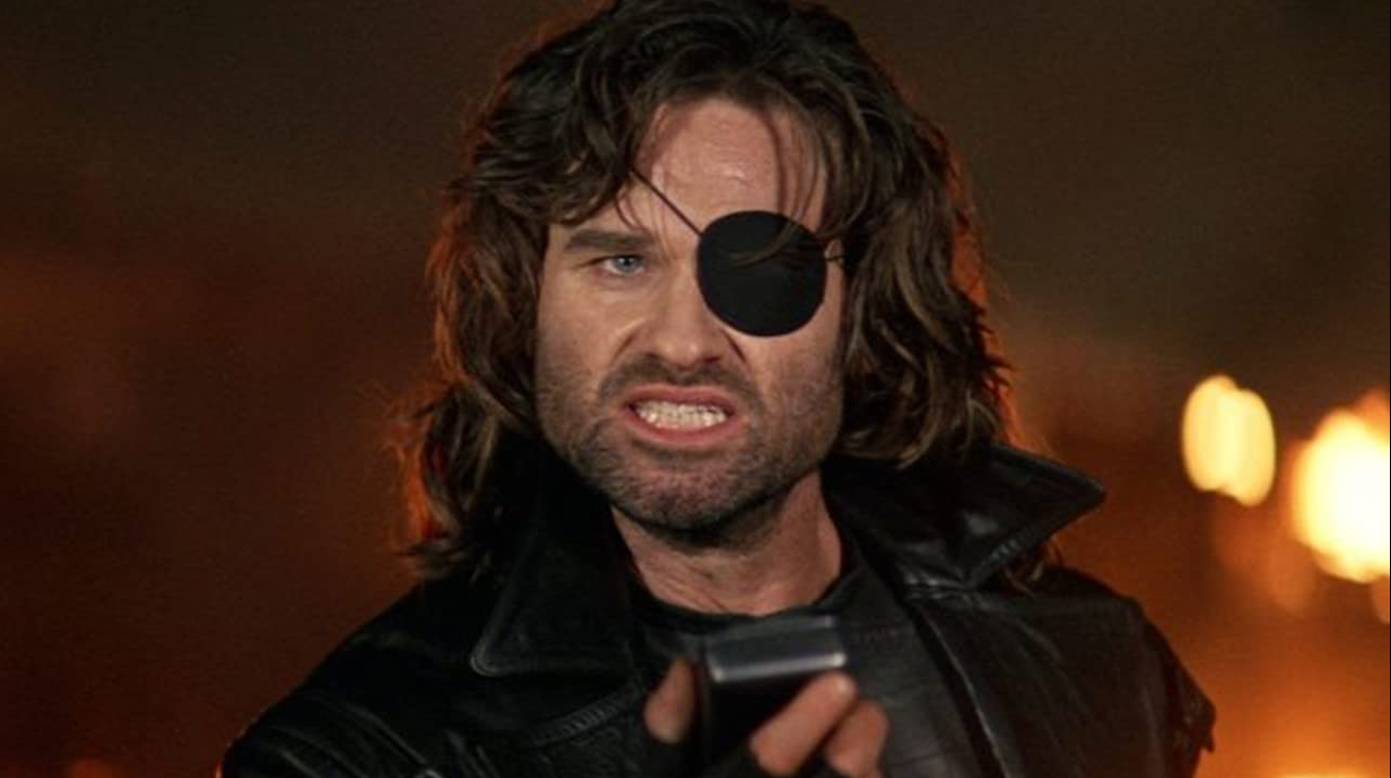 A scowling, bearded man played by actor Kurt Russell with black eye patch talking into a recorder