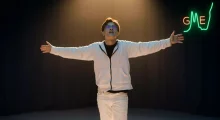A young white man in a white jacket and pants, hands outstretched, under a spotlight on an empty stage