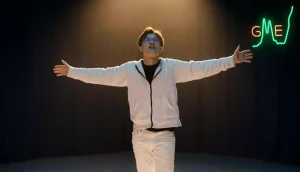 A young white man in a white jacket and pants, hands outstretched, under a spotlight on an empty stage