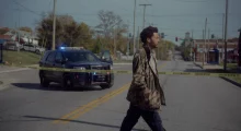 A Black man in a green jacket walking across the street with a police car passing by