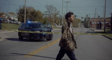 A Black man in a green jacket walking across the street with a police car passing by