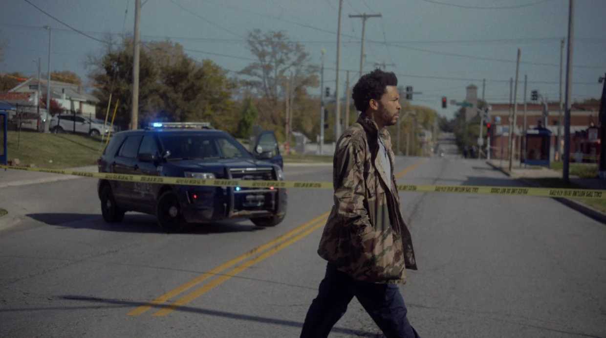 A Black man in a green jacket walking across the street with a police car passing by