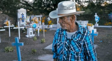 An older Mexican-American man in white cowboy hat and blue-checked shirt in a graveyard