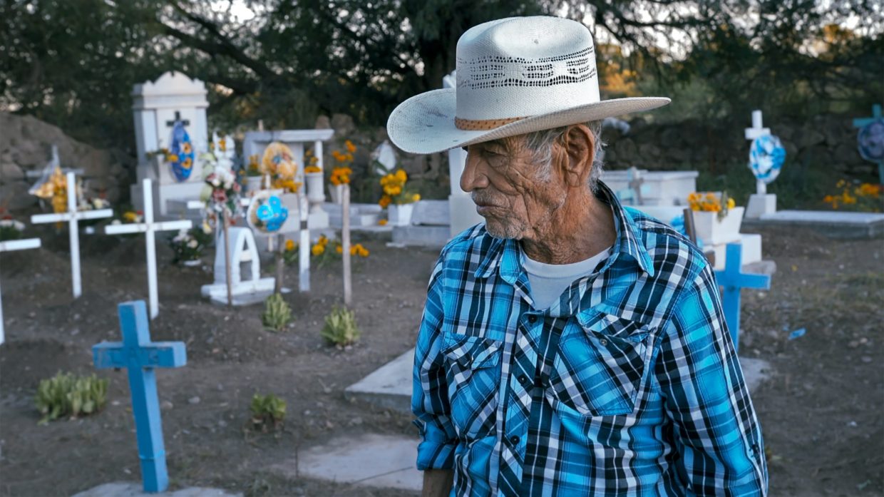 An older Mexican-American man in white cowboy hat and blue-checked shirt in a graveyard