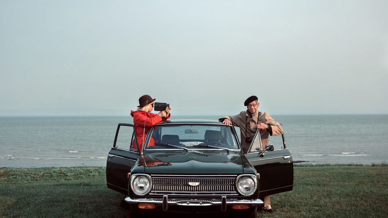 A car on a beach with the sea in the background. The doors are open, and a woman in a red top is from one side filming a man in a beret on the other