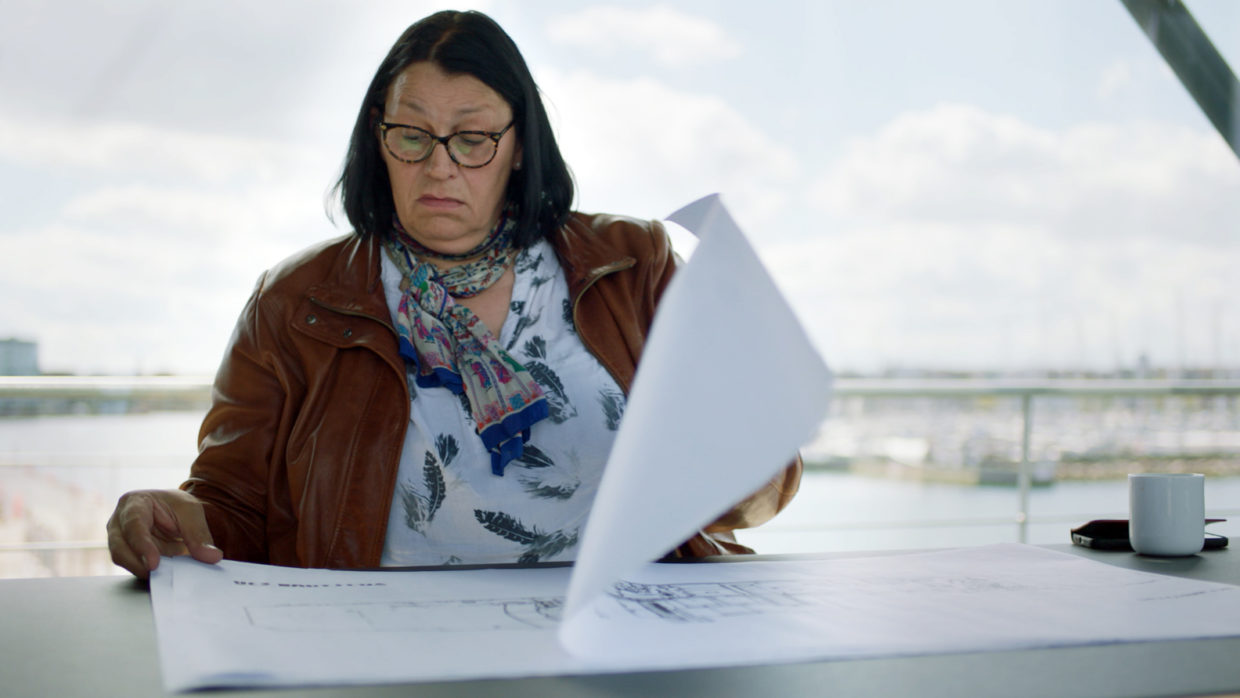 A middle-aged woman with brown hair wearing a red jacket looking at papers in a scene from the documentary "Undercurrent: The Disappearance of Kim Wall"