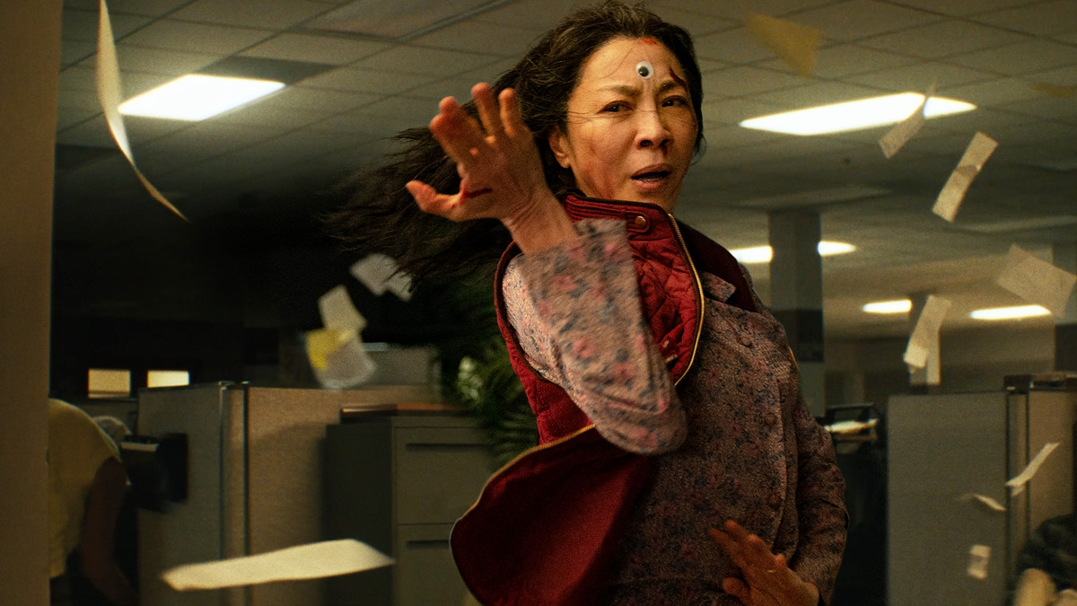 an Asian woman with long black hair standing, arm outstretched, amidst a swirl of papers in an office