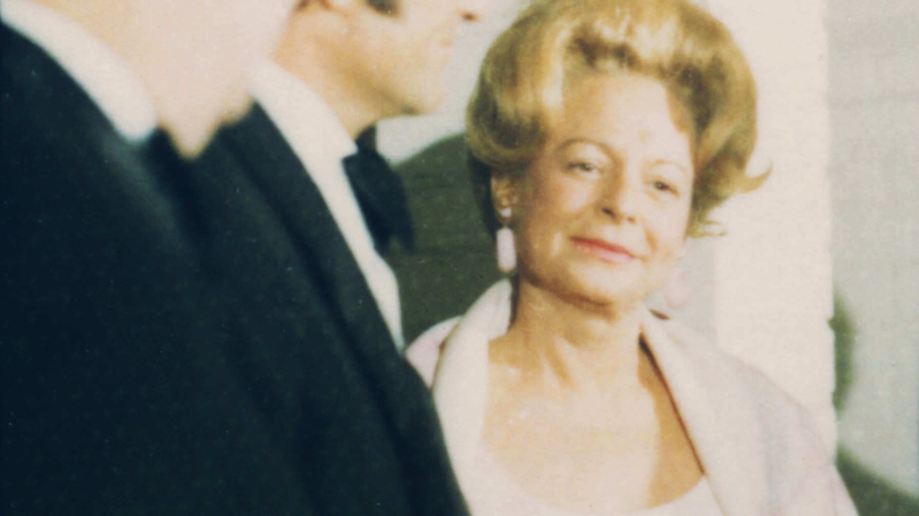 An older white woman with blonde hair and an open necked blouse with two men in suits