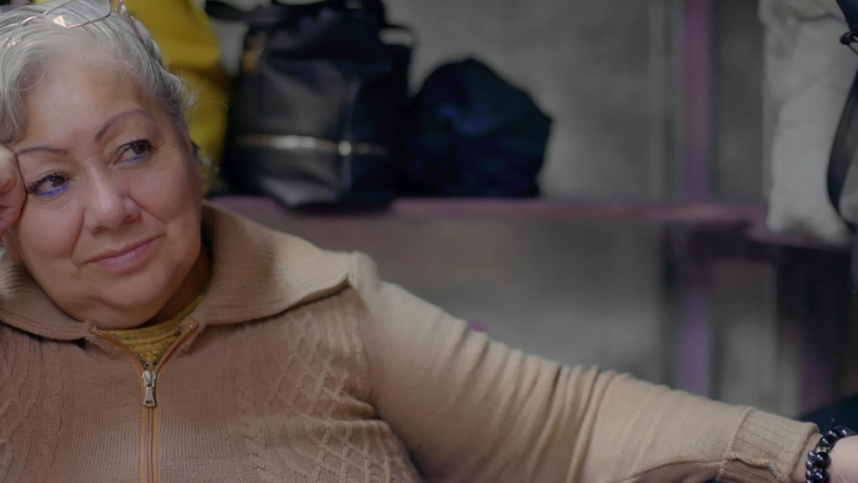 An older Mexican woman with a beige top and gray hair reclining on a sofa