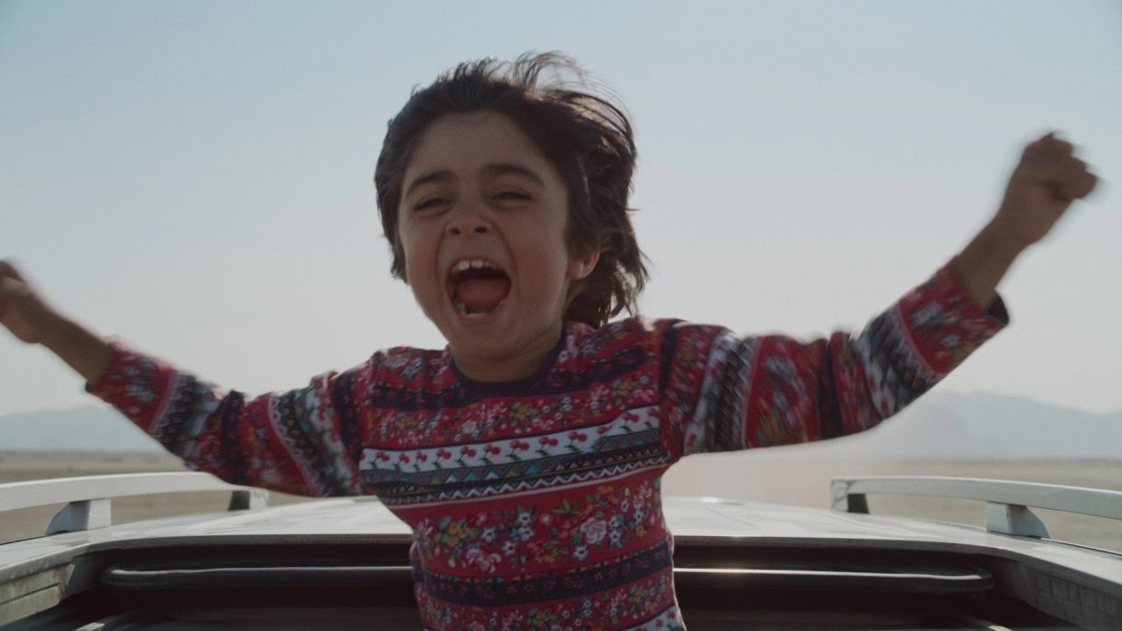 A young Iranian boy in a multicolored sweater with arms outstretched as he juts out of the roof of an automobile