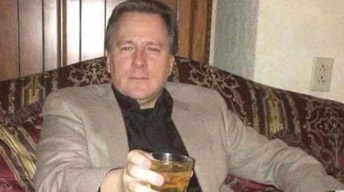 A middle-aged white man in a black shirt and tan jacket holding a drink