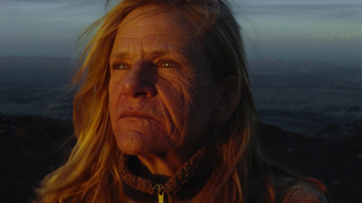 A middle-aged woman with sun-parched skin and blonde hair outdoors looking into the distance