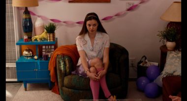 A young woman with brown hair wearing a light-colored patterned top and pink stockings, sitting in a brown chair