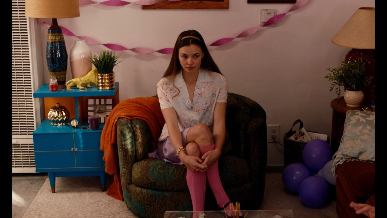 A young woman with brown hair wearing a light-colored patterned top and pink stockings, sitting in a brown chair