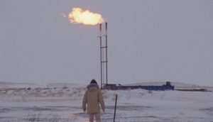 A figure in a heavy winter coat stands in front of an oil pipeline shrouded in fresh snowfall.