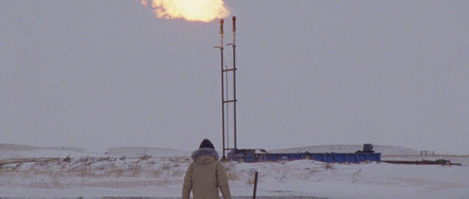 A figure in a heavy winter coat stands in front of an oil pipeline shrouded in fresh snowfall.