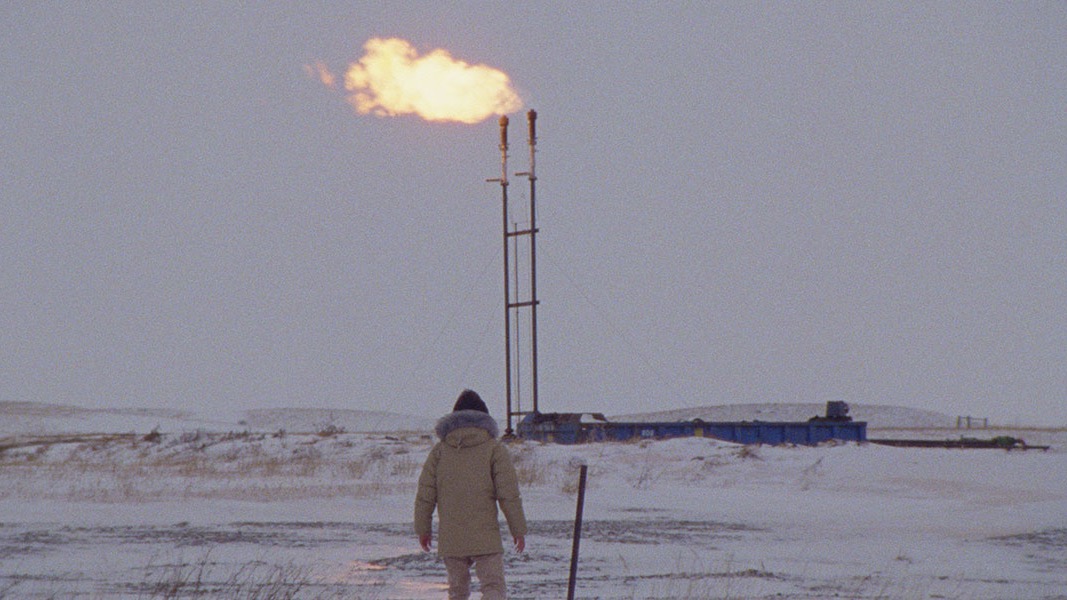 A figure in a heavy winter coat stands in front of an oil pipeline shrouded in fresh snowfall.