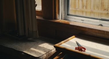 A pair of scissors with a red handle and lying on a table illuminated by sunlight coming in through a window.