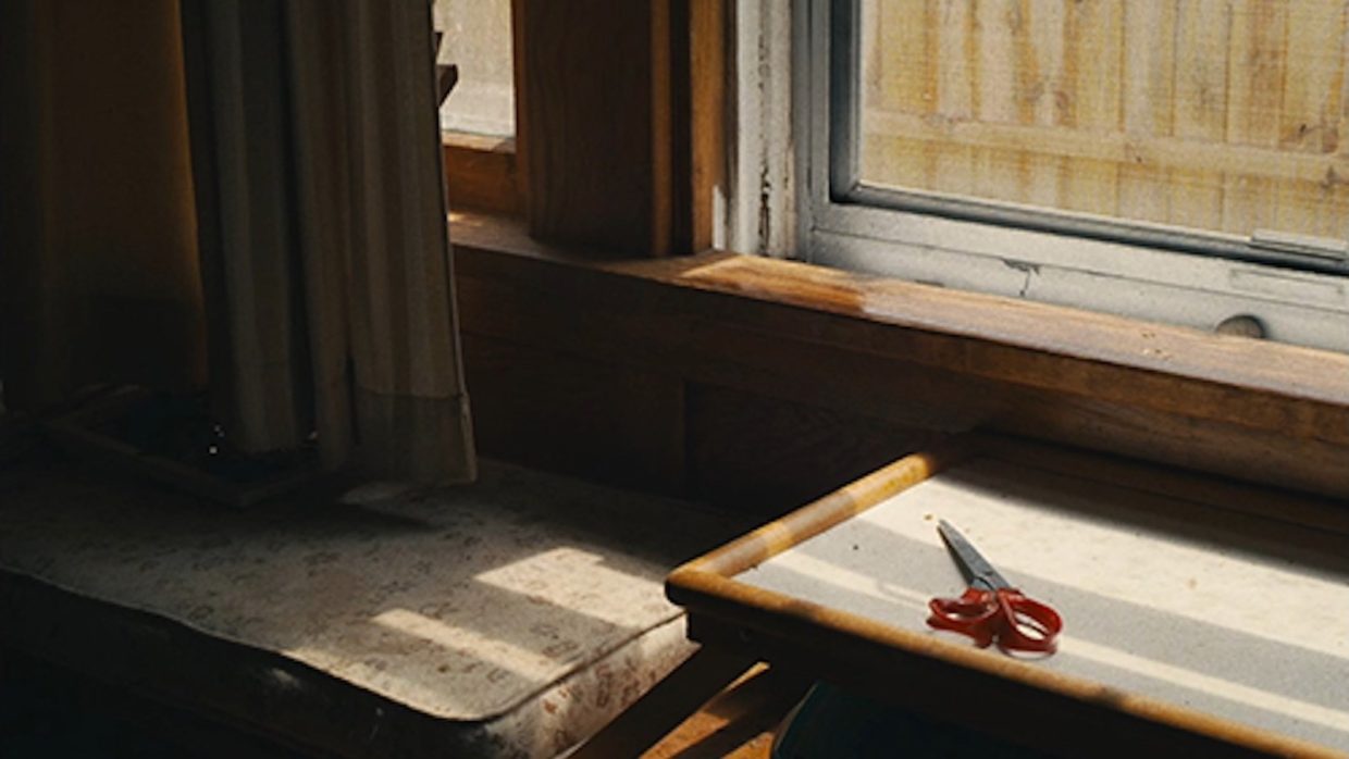 A pair of scissors with a red handle lying on a table and lit by sunlight coming through a window.