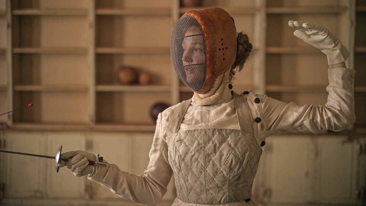 A woman wears her brown hair in a bun behind her head, she also wears a fencing uniform, mask and holds a sword.