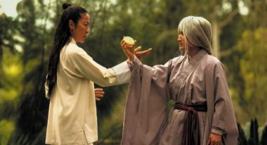 Michelle Yeoh wears a pale yellow smock with half of her long, black hair pulled into a bun. Her hand touches Ke Huy Quan's wrist. Quan dons a gray wrobe with long bell sleeves and a long gray wig. In his outstretched hand, he holds a cookie.
