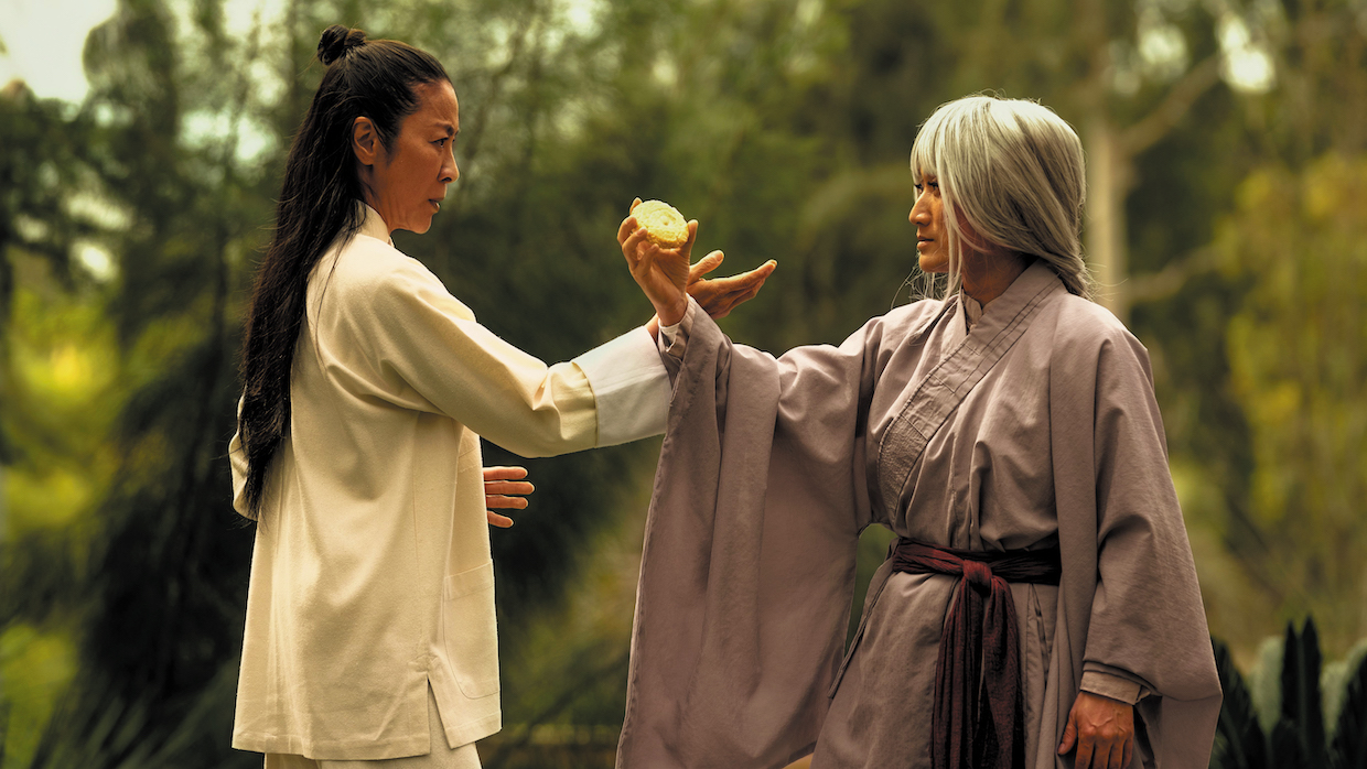 Michelle Yeoh wears a pale yellow smock with half of her long, black hair pulled into a bun. Her hand touches Ke Huy Quan's wrist. Quan dons a gray wrobe with long bell sleeves and a long gray wig. In his outstretched hand, he holds a cookie.