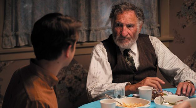 The back of a young boy's head with brown hair is turned toward an elderly man wearing a white shirt with pinstripes and a brown sweater vest. They are sitting at a dining table during dinnertime.