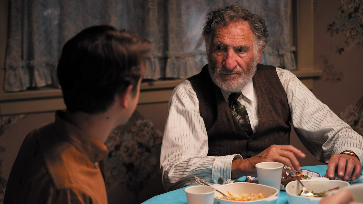 The back of a young boy's head with brown hair is turned toward an elderly man wearing a white shirt with pinstripes and a brown sweater vest. They are sitting at a dining table during dinnertime.