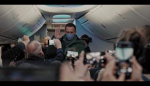 A masked Alexei Navalny waves as he boards a plane while passengers take photos and videos of him.