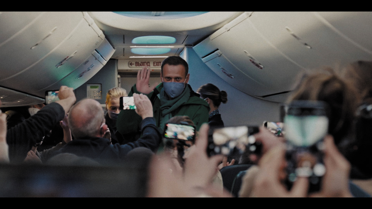 A masked Alexei Navalny waves as he boards a plane while passengers take photos and videos of him.