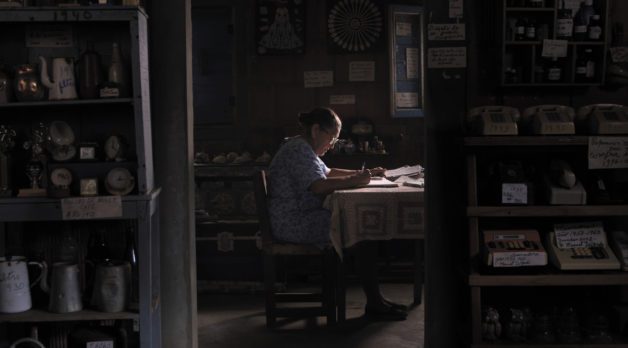 A woman in a blue floral house dress sits at a wooden dining table with a pen and paper, surrounded by curated clutter in wood-paneled room.