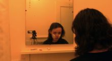 A teenage girl with cropped brown hair stares into the bathroom mirror, a handheld camera is place on a tripod right next to her. She has white paint on her cheeks.