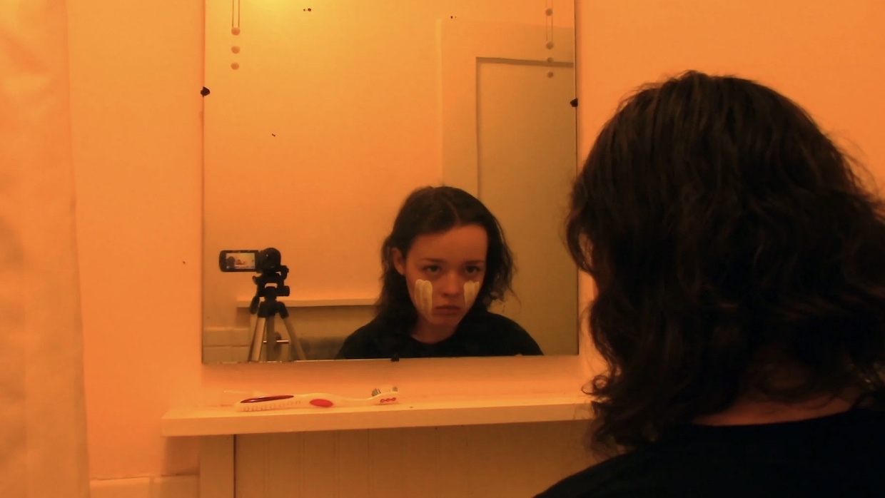 A teenage girl with cropped brown hair stares into the bathroom mirror, a handheld camera is place on a tripod right next to her. She has white paint on her cheeks.