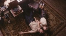 Two lovers circa the 1940s lie on a rug next to a wooden office char, desk and record player. The woman holds the bespectacled man's face in her hand.