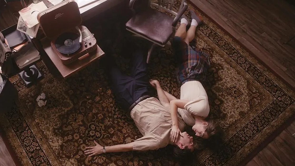 Two lovers circa the 1940s lie on a rug next to a wooden office char, desk and record player. The woman holds the bespectacled man's face in her hand.