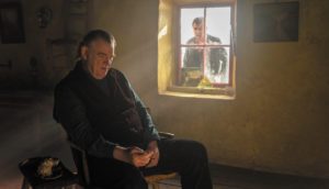 An elderly Irish man named Colm (Brendan Gleeson) sits in a chair next to a window. His younger friend Pádraic (Colin Farrell) stares at him through the window. They do not make eye contact.