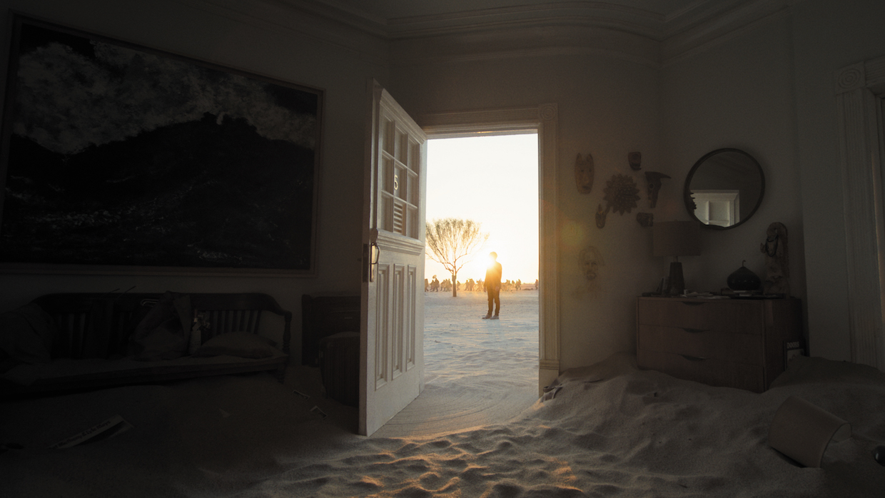 Inside of a house filled with sand, the front door is wide open, revealing the shadow of a man standing far off in the background.