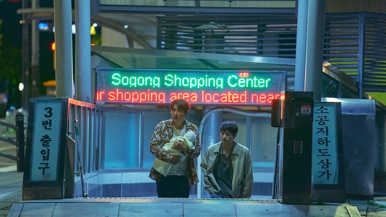 Two Korean men ascend an escalator in a shopping center, with the older of the two men holding a baby in his arms.