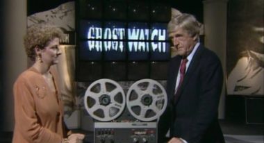 A woman wearing an orange blazer with short, curly, blond hair stands opposite a white man with gray hair and a blue suit in a TV studio. Projected behind them is the logo for "Ghostwatch," and they stand next to a '90s-era audio player.